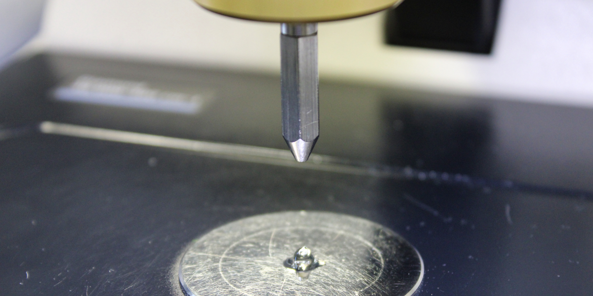 Auf einer Metallplatte ist eine Tupfer eine cremigen Substanz aufgebracht, darüber schwebt ein Metallstift.
