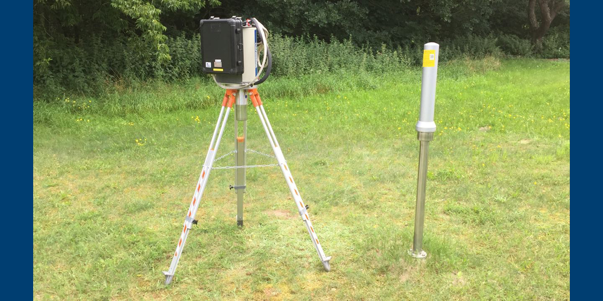 Bild: Auf einer Wiese steht ein Messgerät auf einem dreibeinigen Stativ neben einem in der Wiese verankerten schmalen Metallzylinder.