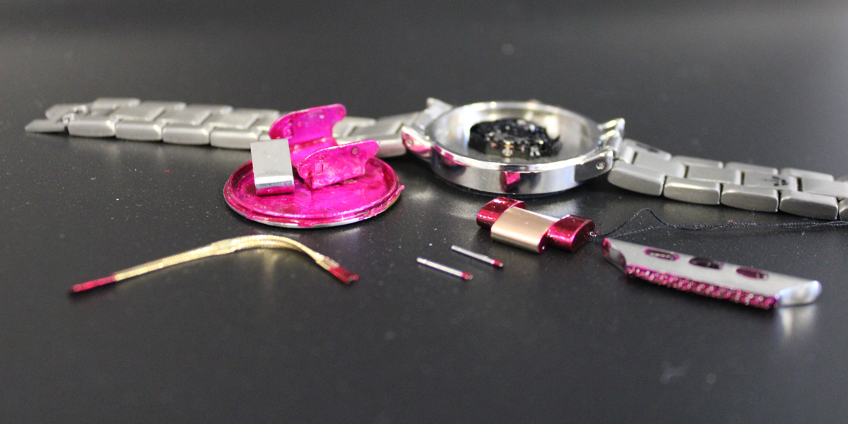 Bild: Eine in Einzelteile zerlegte Armbanduhr aus Metall. Teile, die beim Tragen der Uhr nicht mit der Haut in Berührung kommen, sind rot lackiert.