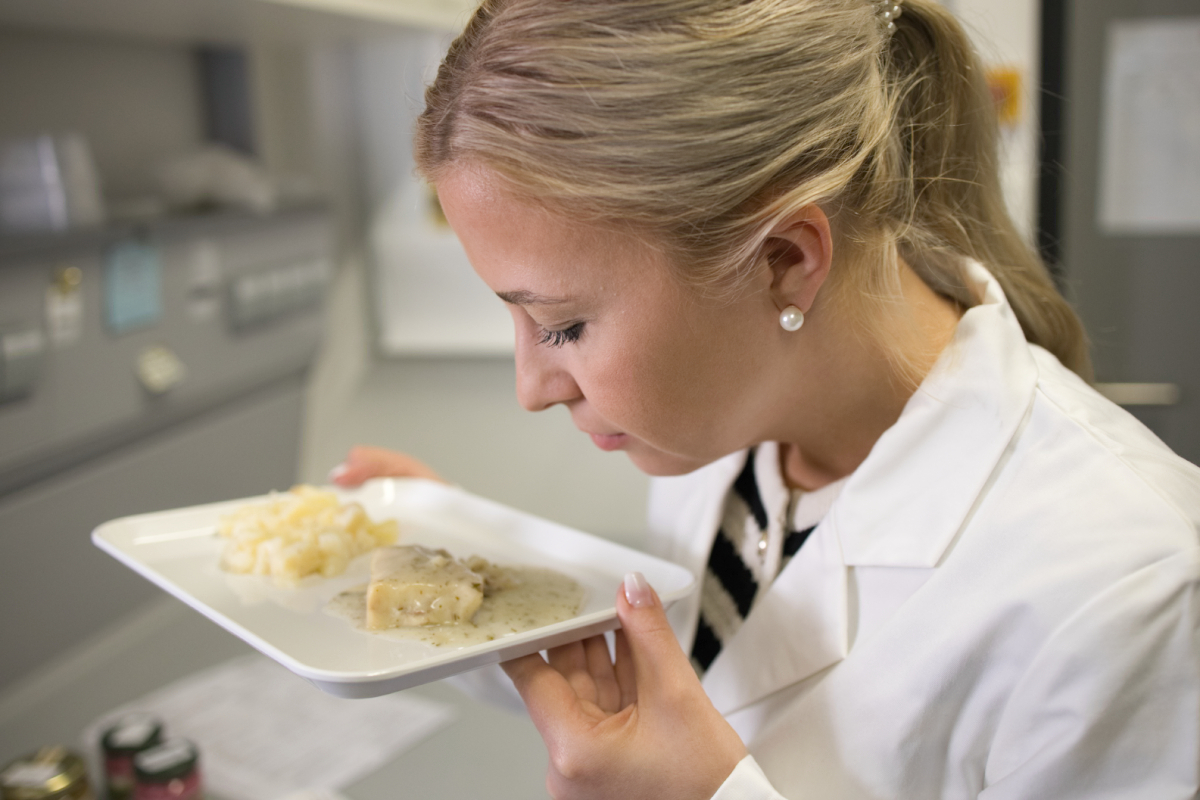 Eine Labormitarbeiterin riecht an einem zubereiteten Fisch-Feriggericht, das auf einer rechteckigen weißen Platte angerichtet ist.