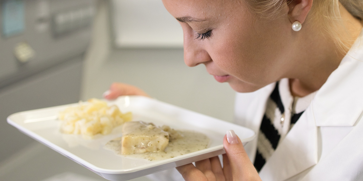 Eine Labormitarbeiterin riecht an einem zubereiteten Fisch-Feriggericht, das auf einer rechteckigen weißen Platte angerichtet ist.