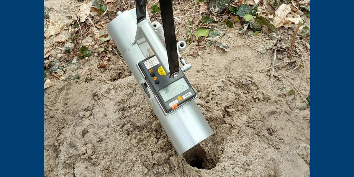Bild: Ein metallfarbenes, zylindrisches, etwa 30 cm langes Ortdosisleistungsmessgerät mit Anzeige wird über ein Bohrloch im Sandboden gehalten.