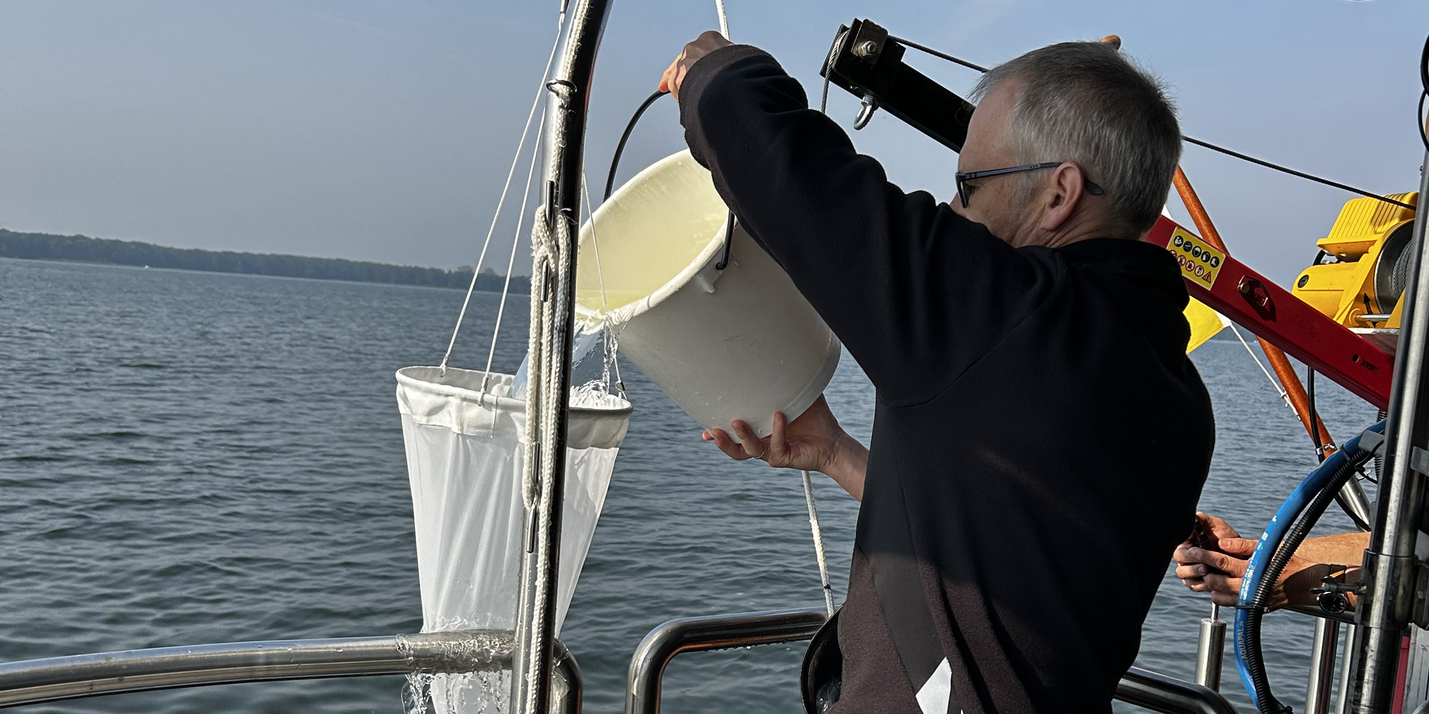 Ein Mann auf einem Boot gießt Wasser aus einem Eimer durch ein längliches, engmaschiges Netz, das an der Reling über dem Wasser aufgehängt ist.